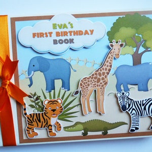 Puede incluir: Un &aacute;lbum de recortes hecho a mano titulado "Eva's First Birthday Book" con tem&aacute;tica de safari. La portada presenta ilustraciones de una jirafa, una cebra, un tigre, un elefante, un rinoceronte y un cocodrilo. Una cinta naranja est&aacute; atada en el lado izquierdo.