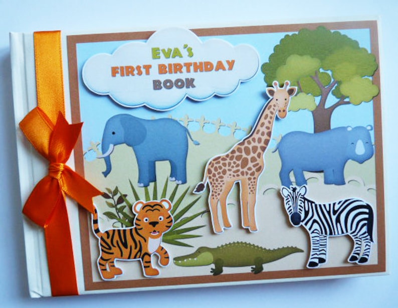 Puede incluir: Un &aacute;lbum de recortes hecho a mano titulado "Eva's First Birthday Book" con tem&aacute;tica de safari. La portada presenta ilustraciones de una jirafa, una cebra, un tigre, un elefante, un rinoceronte y un cocodrilo. Una cinta naranja est&aacute; atada en el lado izquierdo.