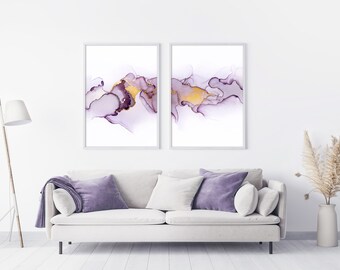 Puede incluir: Dos impresiones de arte abstracto enmarcadas con fondo blanco y diseños de acuarela en tonos morados y dorados. Las impresiones están colgadas en una pared blanca sobre un sofá blanco con almohadas moradas.