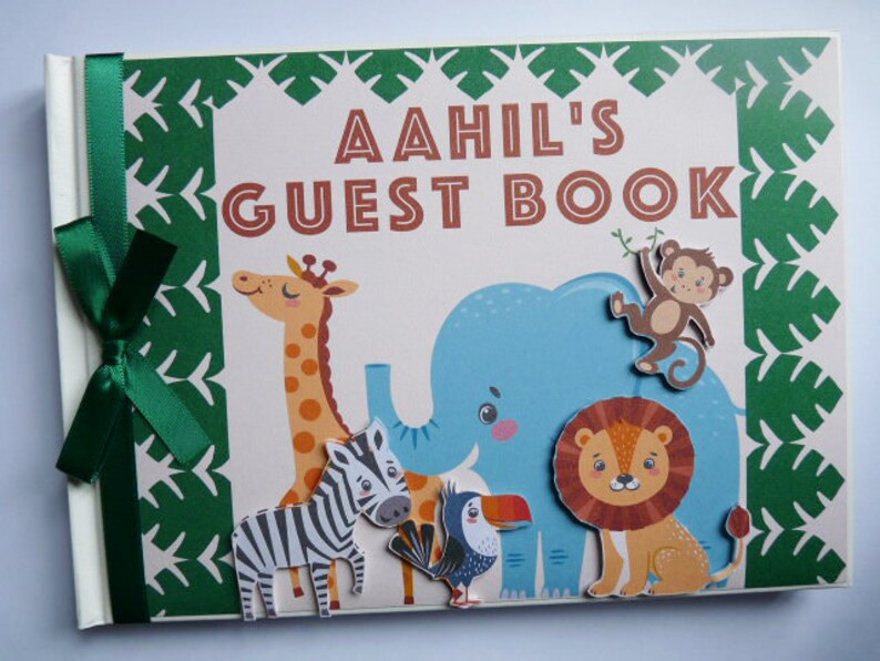 Puede incluir: Un libro de visitas personalizado con el nombre "Aahil" en letras marrones. La portada presenta un tema de jungla verde y blanco con ilustraciones de una jirafa, una cebra, un elefante, un mono, un le&oacute;n y un tuc&aacute;n. Una cinta verde est&aacute; atada en el lomo.