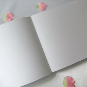 May include: An open, blank sketchbook with white pages. The book is lying open, with the pages angled towards the viewer. Scattered around the book are pink and green flower petals, adding a touch of color and softness to the scene.
