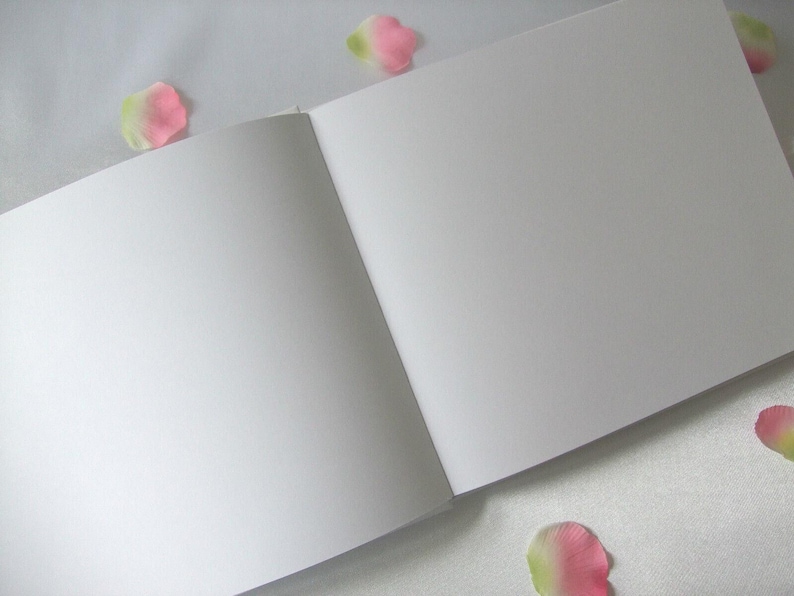 May include: An open, blank sketchbook with white pages. The book is lying open, with the pages angled towards the viewer. Scattered around the book are pink and green flower petals, adding a touch of color and softness to the scene.