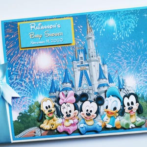 May include: A baby shower scrapbook with a blue cover featuring Disney characters and fireworks. The cover includes the text "Rhiannon's Baby Shower November 14, 2020". A light blue ribbon and bow are attached to the spine.