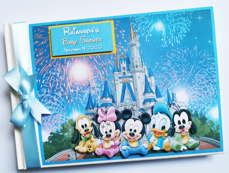 May include: A baby shower scrapbook with a blue cover featuring Disney characters and fireworks. The cover includes the text "Rhiannon's Baby Shower November 14, 2020". A light blue ribbon and bow are attached to the spine.