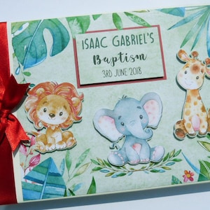 Puede incluir: Un &aacute;lbum de fotos con tem&aacute;tica de bautizo con una cinta y un lazo rojos. La portada presenta ilustraciones de un le&oacute;n, un elefante y una jirafa, junto con el texto "ISAAC GABRIEL'S Baptism 3RD JUNE 2018" sobre un fondo verde claro.