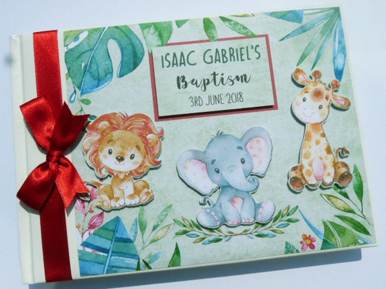 Puede incluir: Un &aacute;lbum de fotos con tem&aacute;tica de bautizo con una cinta y un lazo rojos. La portada presenta ilustraciones de un le&oacute;n, un elefante y una jirafa, junto con el texto "ISAAC GABRIEL'S Baptism 3RD JUNE 2018" sobre un fondo verde claro.