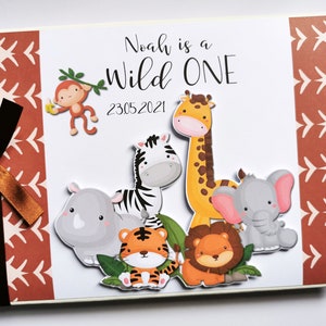 Puede incluir: Un &aacute;lbum de recortes personalizado con un tema de la jungla. La portada presenta animales de dibujos animados, incluyendo una jirafa, una cebra, un le&oacute;n y un elefante. El texto "Noah is a Wild One" se muestra encima de la fecha 23.05.2021. Una cinta marr&oacute;n est&aacute; atada en el lado izquierdo.