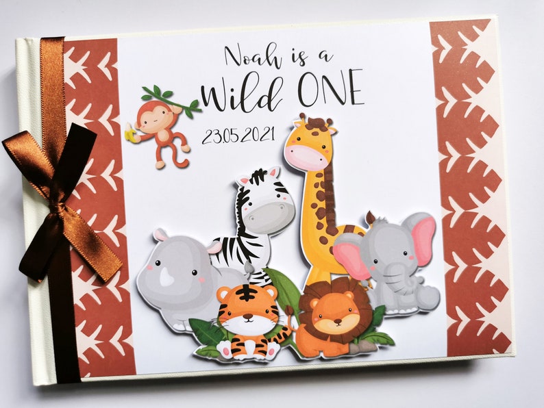 Puede incluir: Un &aacute;lbum de recortes personalizado con un tema de la jungla. La portada presenta animales de dibujos animados, incluyendo una jirafa, una cebra, un le&oacute;n y un elefante. El texto "Noah is a Wild One" se muestra encima de la fecha 23.05.2021. Una cinta marr&oacute;n est&aacute; atada en el lado izquierdo.