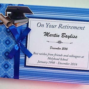May include: A blue and white retirement scrapbook with a blue ribbon and a graduation cap on the cover. The text on the cover reads "On Your Retirement Martin Bayliss December 2016 Best wishes from friends and colleagues at Holyhead School January 1998 - December 2016"
