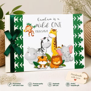 Puede incluir: Un libro de recuerdos personalizado con una cinta verde. La portada muestra el texto "CamCam is a Wild ONE" e ilustraciones de animales de la selva, como un mono, una cebra, una jirafa, un tigre, un le&oacute;n, un rinoceronte y un elefante.