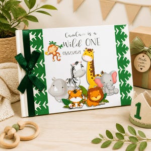 Puede incluir: Un &aacute;lbum de fotos blanco con una cinta verde e ilustraciones de animales de la jungla. El &aacute;lbum dice "CamCam is a Wild One" con la fecha 08/12/2021. La portada presenta un mono, una cebra, una jirafa, un rinoceronte, un tigre, un le&oacute;n y un elefante.