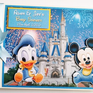 May include: A baby shower memory book with a blue ribbon and bow. The cover features Donald Duck and Mickey Mouse in front of a castle with fireworks. The text on the cover reads "Adam & Joe's Baby Shower 25th April 2020."