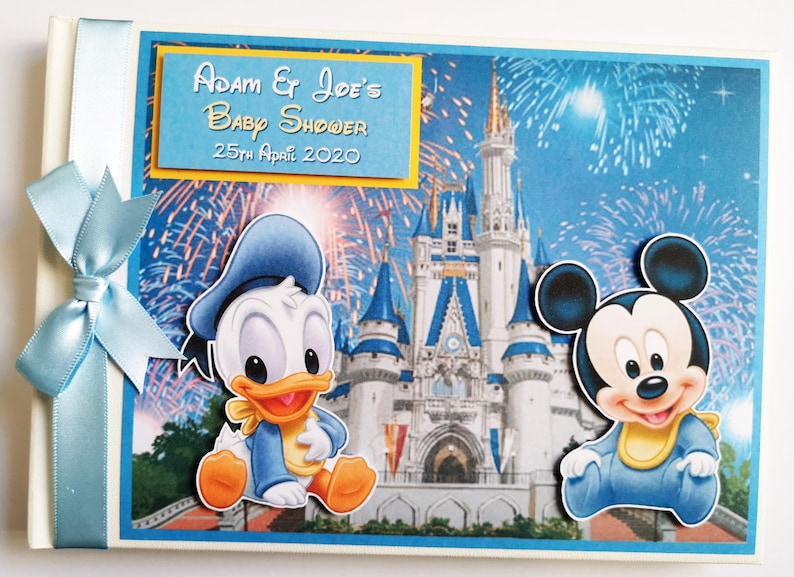 May include: A baby shower memory book with a blue ribbon and bow. The cover features Donald Duck and Mickey Mouse in front of a castle with fireworks. The text on the cover reads "Adam & Joe's Baby Shower 25th April 2020."
