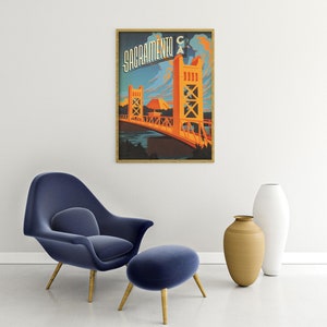 May include: A vintage travel poster featuring a stylised illustration of the Tower Bridge in Sacramento, California. The poster is in a wooden frame and has a blue, orange and yellow colour scheme. The text "Sacramento CA" is printed in a bold, stylised font at the top of the poster.