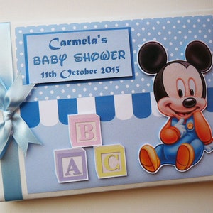 May include: A baby shower invitation with a blue and white polka dot and striped design. The invitation features a Mickey Mouse graphic and the text "Carmela's BABY SHOWER 11th October 2015". A light blue ribbon and bow are tied on the left side.