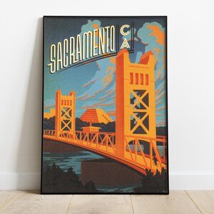 May include: A vintage-style poster featuring a stylised illustration of the Tower Bridge in Sacramento, California. The poster is in a black frame and has a blue, orange, and yellow colour scheme. The text "Sacramento CA" is written in a stylised font at the top of the poster.