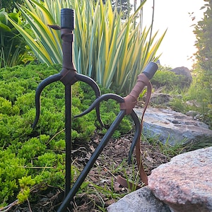 May include: Two black sai, traditional Japanese martial arts weapons, are displayed outdoors. The sai have a central shaft with a handle wrapped in brown leather and two curved prongs. The background features green foliage, rocks, and flowers.