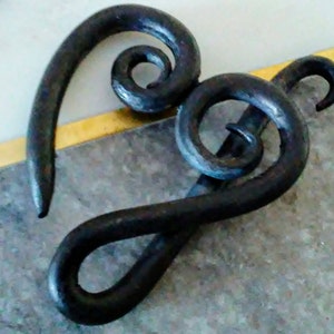 May include: A handcrafted, dark metal decorative hook with a unique, swirling design. The hook features a series of curved and looped elements, creating an artistic and functional piece. The metal has a textured surface and a dark, almost black color.