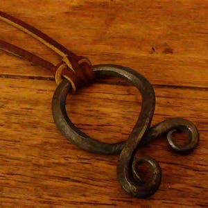 May include: A brown leather cord necklace with a hand-forged metal pendant. The pendant is a twisted circle with a smaller circle at the bottom.