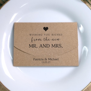 Peut inclure: Une enveloppe rectangulaire en papier kraft brun avec un rabat, ornée d'une icône de cœur noir et du texte imprimé : WISHING YOU RICHES from the new MR. AND MRS. Patricia & Michael 13.05.27. L'enveloppe est centrée sur une assiette blanche.