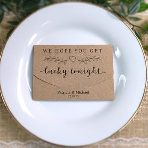 May include: A brown card with the words "WE HOPE YOU GET lucky tonight" in cursive, with a heart and floral design. The card sits on a white plate with a gold edge, ideal for wedding or event decorations. The names Patricia & Michael and the date are also visible.