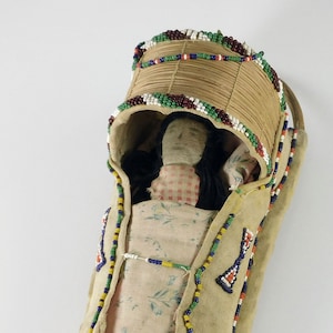 May include: A beaded cradle board with a doll inside. The cradle board is made of wood and is decorated with colorful beads. The doll is wearing a pink and white dress and has black hair.