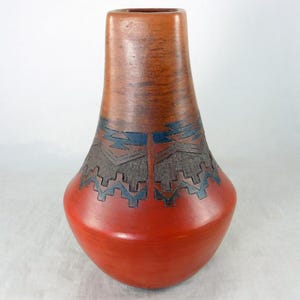 May include: A handcrafted ceramic vase with a distinctive design. The vase features a vibrant red base, transitioning to a brown upper section. Intricate geometric patterns in black and blue adorn the midsection, creating a striking contrast. The vase has a rounded body and a narrow neck.
