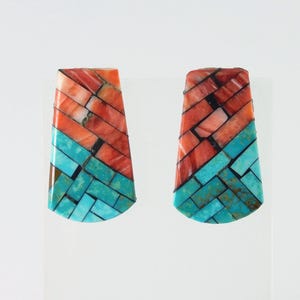 May include: A pair of handcrafted earrings with a geometric design. The earrings feature a combination of vibrant turquoise and orange-red stones, arranged in a brick-like pattern. The stones are set in a silver or metal base, creating a striking contrast.