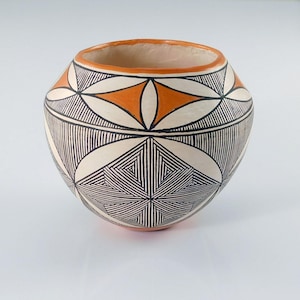 May include: A round ceramic pot featuring a cream-colored base with intricate black line patterns and orange geometric accents. The vessel has a wide opening with a smooth orange rim and a rounded body decorated with repeating star-like motifs.