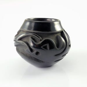 May include: A black ceramic pot with a rounded body and a wide opening. The pot features carved, abstract designs around its exterior. The surface has a glossy finish, and the pot sits on a white surface.