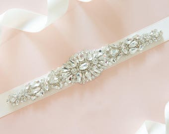 May include: A bridal sash with a central cluster of clear crystals and rhinestones, flanked by smaller crystal accents, set on a wide white satin ribbon. The ribbon is displayed on a soft pink background with white ribbon accents.