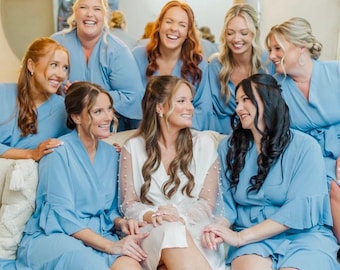 May include: A group of women in a well-lit room, smiling and laughing. Most are wearing light blue robes with ruffled sleeves. One woman is wearing a white robe with pearl accents. The women are sitting on a beige couch in front of oval mirrors.