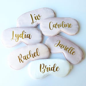 May include: A collection of sleep masks in various shades of pink and white. Each mask is personalized with a name in gold script, including "Liv", "Caroline", "Lydia", "Rachel", and "Janelle". One mask is white and says "Bride".