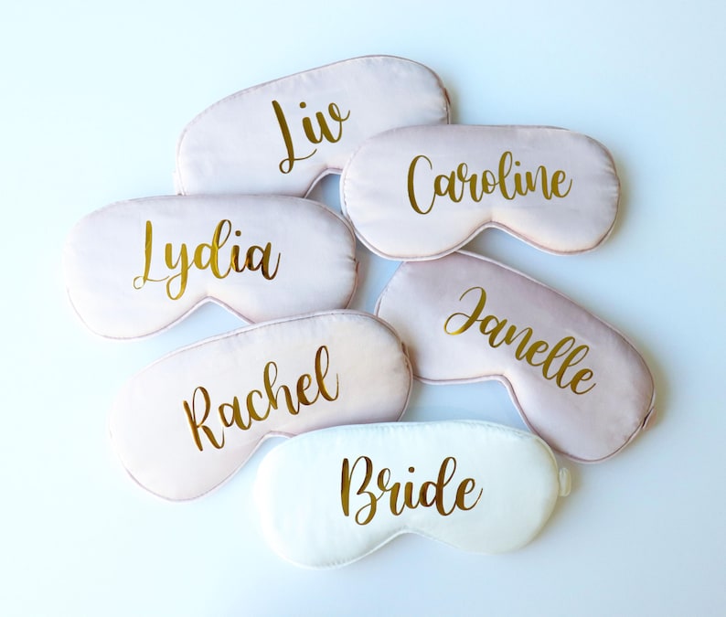 May include: A collection of sleep masks in various shades of pink and white. Each mask is personalized with a name in gold script, including "Liv", "Caroline", "Lydia", "Rachel", and "Janelle". One mask is white and says "Bride".