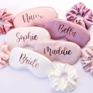 May include: A collection of satin sleep masks and matching scrunchies in shades of pink and white. The sleep masks are personalized with names in a metallic script, including "Naomi", "Sophie", "Bella", "Maddie", and "Bride".