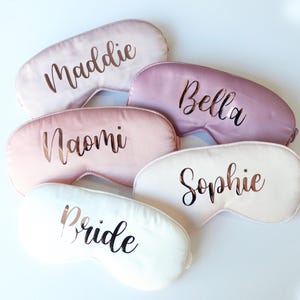 May include: A collection of personalized sleep masks in various pastel colors, including pink and white. Each mask features a name or the word "Bride" in a metallic script font. The masks are arranged on a white surface.