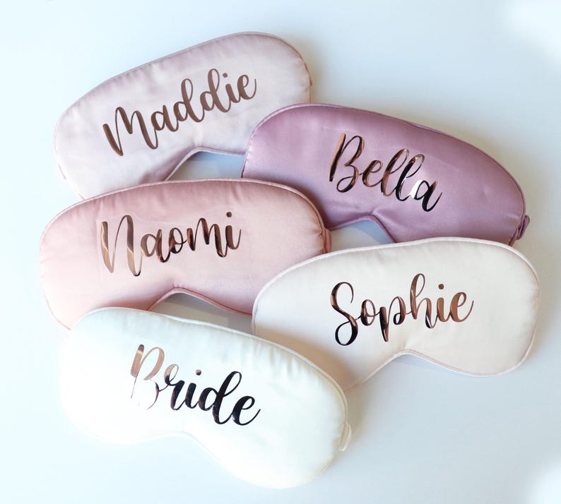 May include: A collection of personalized sleep masks in various pastel colors, including pink and white. Each mask features a name or the word "Bride" in a metallic script font. The masks are arranged on a white surface.