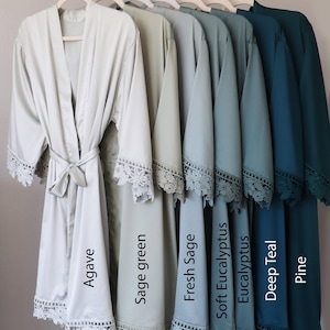 Op de afbeelding: Zeven satijnen badjassen in verschillende tinten groen en blauw, elk met een kantversiering. De badjassen hangen aan hangers en zijn gelabeld met de kleurennamen: Agave, Saliegroen, Verse Salie, Zachte Eucalyptus, Eucalyptus, Diep Teal en Pine.