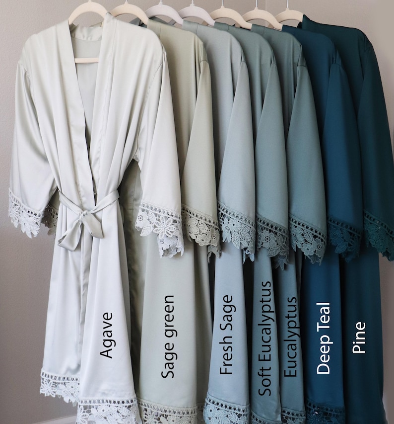 Op de afbeelding: Zeven satijnen badjassen in verschillende tinten groen en blauw, elk met een kantversiering. De badjassen hangen aan hangers en zijn gelabeld met de kleurennamen: Agave, Saliegroen, Verse Salie, Zachte Eucalyptus, Eucalyptus, Diep Teal en Pine.