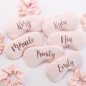 May include: A collection of personalized sleep masks and matching scrunchies in a soft pink hue. Each sleep mask is inscribed with a name in a stylish script font. The scrunchies are made of a silky material.