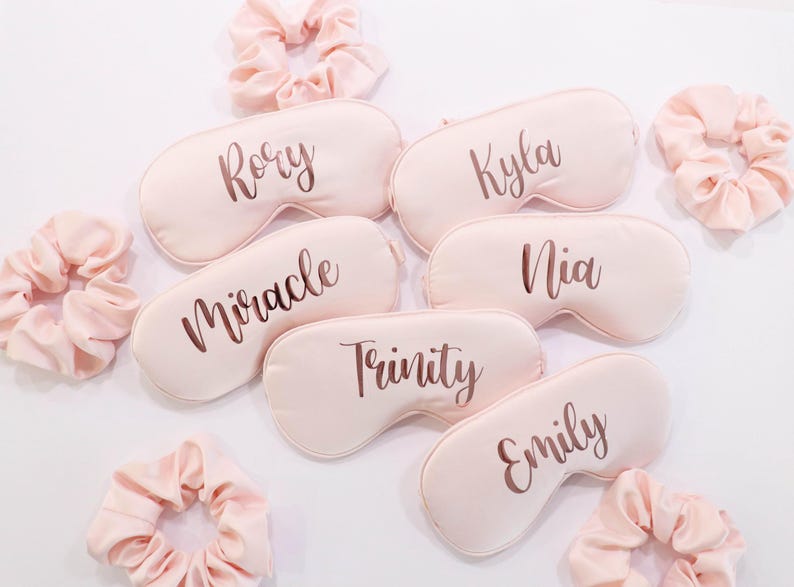 May include: A collection of personalized sleep masks and matching scrunchies in a soft pink hue. Each sleep mask is inscribed with a name in a stylish script font. The scrunchies are made of a silky material.