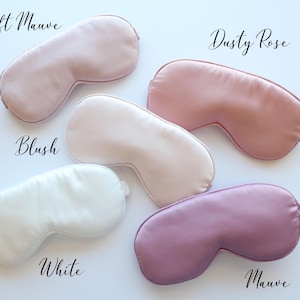 May include: Five satin sleep masks in shades of pink, white, and purple are displayed on a white surface. The masks are labeled "Soft Mauve", "Dusty Rose", "Blush", "White", and "Mauve". The masks are designed to block out light.