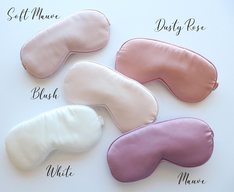 May include: Five satin sleep masks in shades of pink, white, and purple are displayed on a white surface. The masks are labeled "Soft Mauve", "Dusty Rose", "Blush", "White", and "Mauve". The masks are designed to block out light.