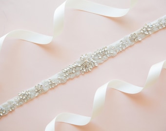 May include: A white satin ribbon with a crystal embellished sash. The sash is decorated with a floral design and features clear rhinestones.