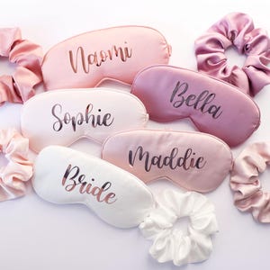 May include: A collection of satin sleep masks and scrunchies in shades of pink and white. The sleep masks are personalized with names like "Naomi", "Sophie", "Bella", "Maddie", and "Bride". The scrunchies are in matching colors.