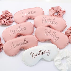 May include: A collection of personalized sleep masks and matching scrunchies. The masks are in shades of pink and white, with names like "Lauren" and "Brittany" printed on them. The scrunchies are made of a silky material.