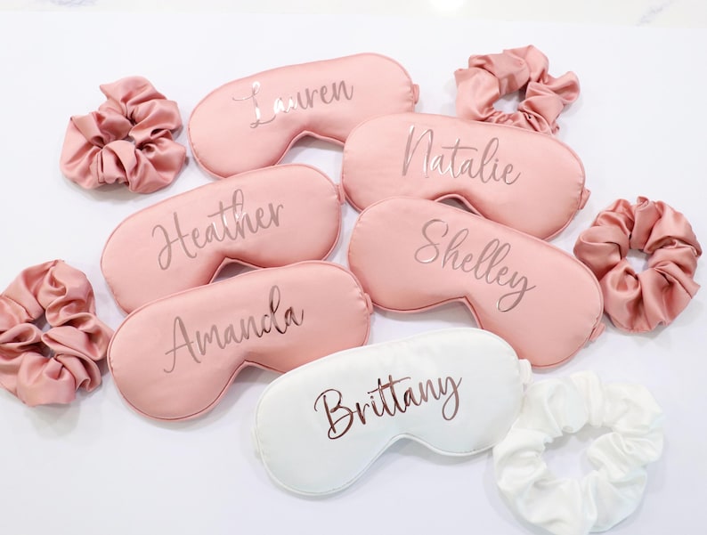 May include: A collection of personalized sleep masks and matching scrunchies. The masks are in shades of pink and white, with names like "Lauren" and "Brittany" printed on them. The scrunchies are made of a silky material.