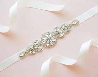 May include: A white satin ribbon with a crystal embellishment. The embellishment is made of clear rhinestones and is shaped like a flower.