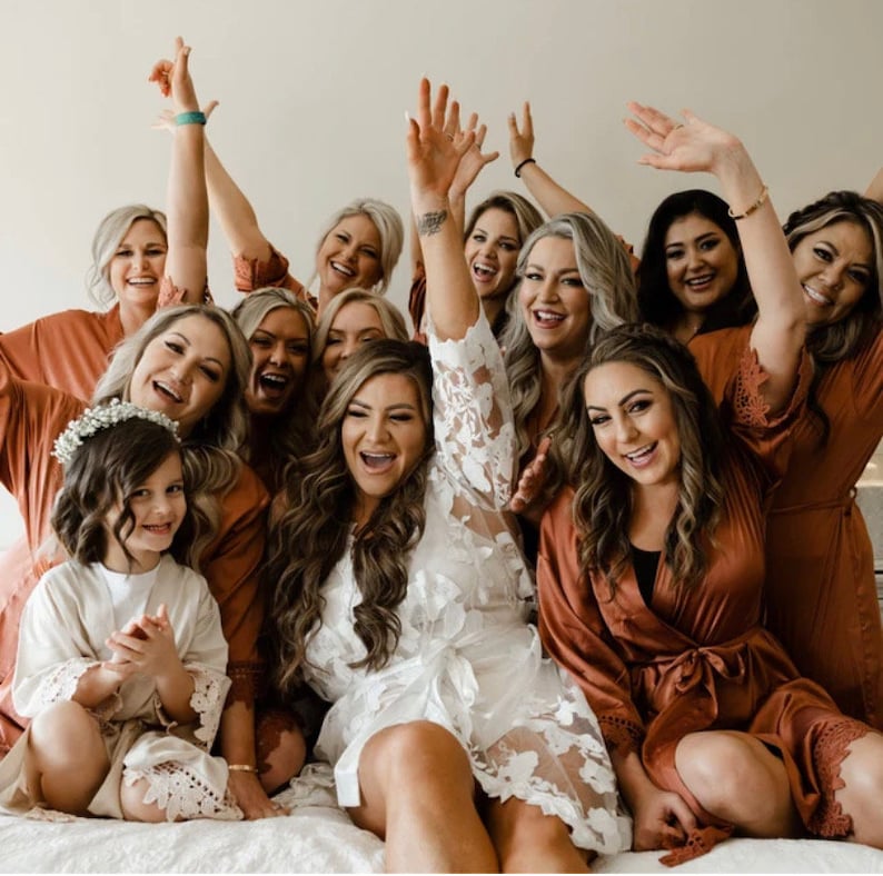 May include: A group of women in matching rust-coloured robes with lace trim, and one woman in a white lace robe, are smiling and laughing. One woman is holding up her hands in the air. A young girl in a white lace robe with a flower crown is sitting at the front.