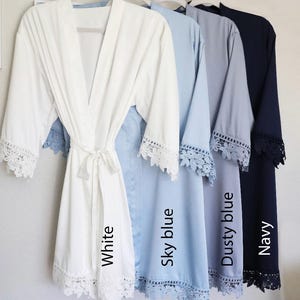 May include: Five satin robes with lace trim in white, sky blue, dusty blue, and navy. The robes are arranged from left to right with the colours listed. The robes have a tie at the waist and are labelled with the colour name.